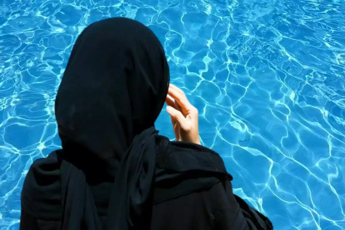 Dona amb hijab negre d’esquena al costat d’una piscina d’aigua blava