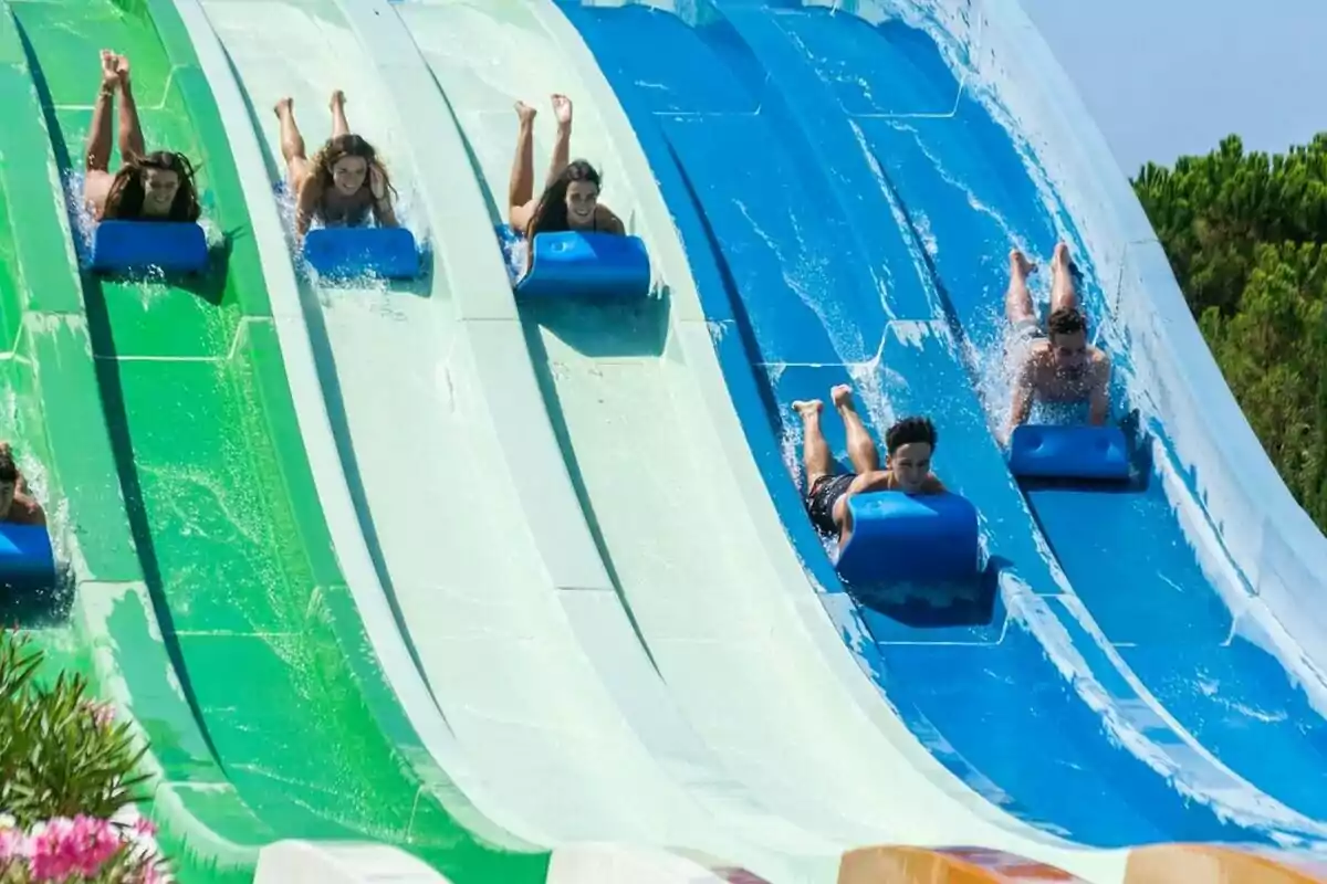 Diverses persones lliscant sobre matalassos per tobogans d’aigua de color verd i blau en un parc aquàtic sota un cel serè Diverses persones lliscant sobre matalassos per tobogans d’aigua de color verd i blau en un parc aquàtic sota un cel serè