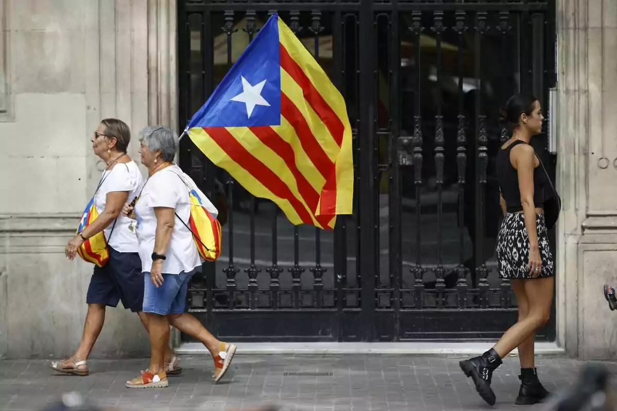 Dues dones grans caminant al costat d'una bandera estelada mentre una dona jove passa pel costat oposat.