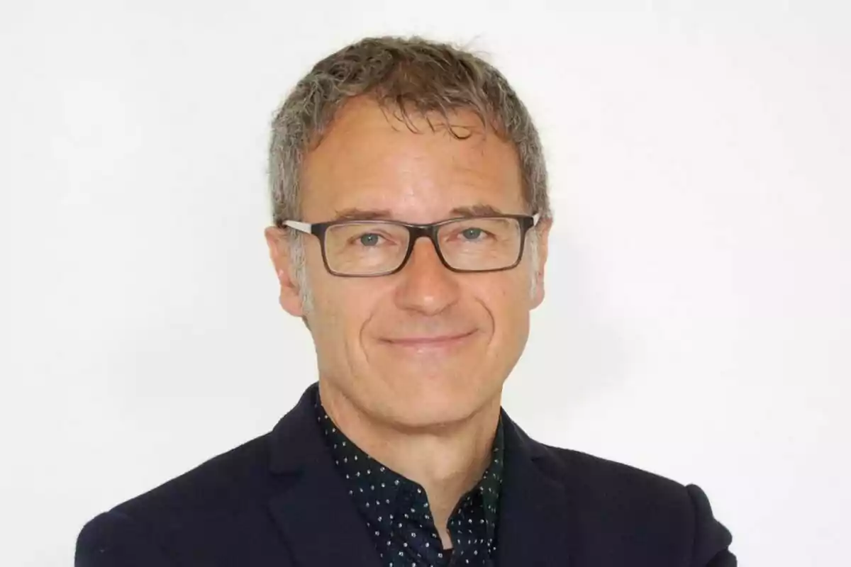 Short-haired, gray-haired man with glasses and a dark jacket smiling in front of a white background Short-haired, gray-haired man with glasses and a dark jacket smiling in front of a white background