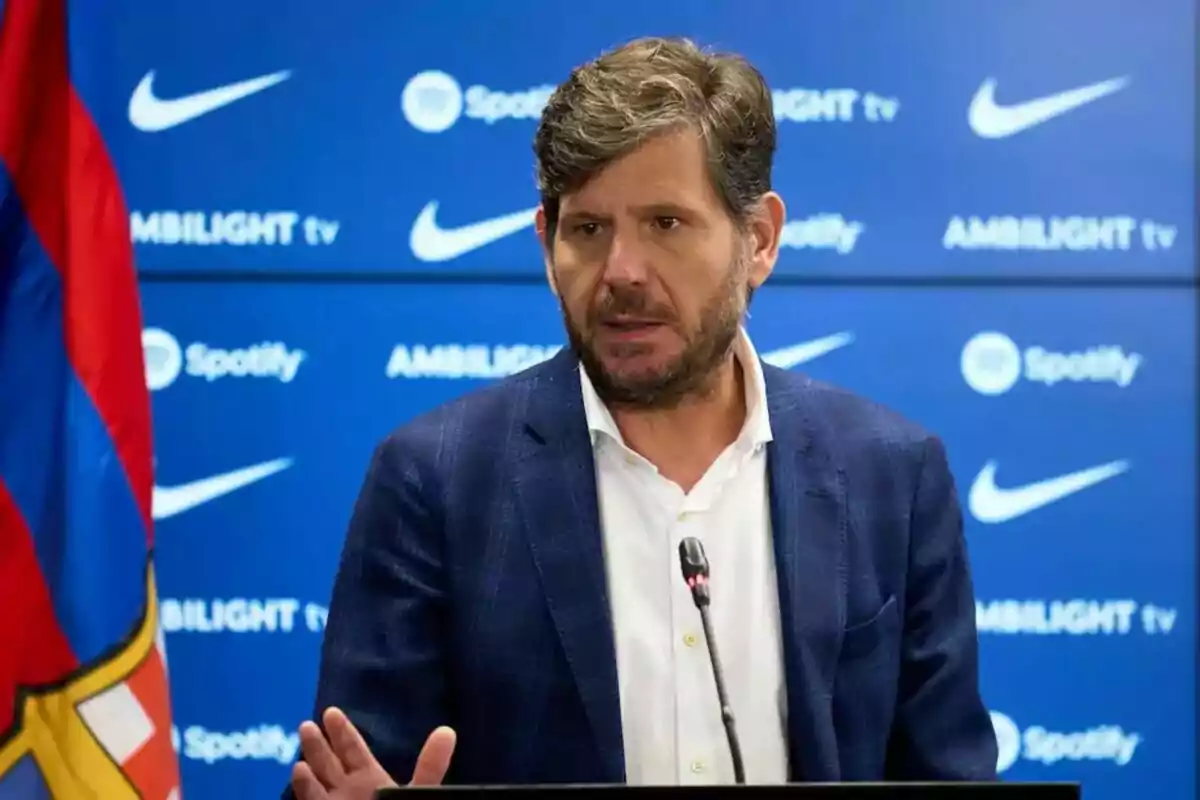 Home amb barba i vestit blau parlant en una conferència de premsa amb un micròfon davant d'un fons blau amb logotips de Nike i Spotify i una bandera a l'esquerra. Home amb barba i vestit blau parlant en una conferència de premsa amb un micròfon davant d'un fons blau amb logotips de Nike i Spotify i una bandera a l'esquerra.