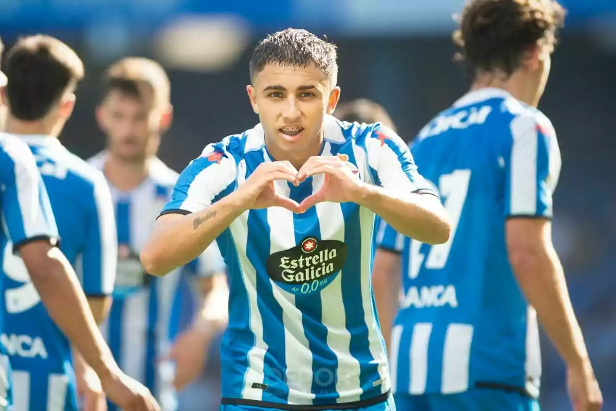 A soccer player in a blue and white striped uniform makes a heart gesture with his hands while other players stand in the background. A soccer player in a blue and white striped uniform makes a heart gesture with his hands while other players stand in the background.