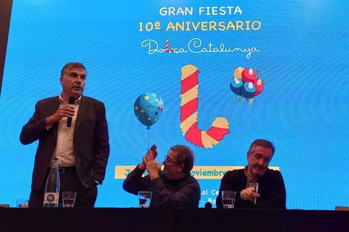 Joan López Alegre dret i Sergio Fidalgo i Pau Guix asseguts i darrere una imatge blava amb el logo de Dolça Catalunya