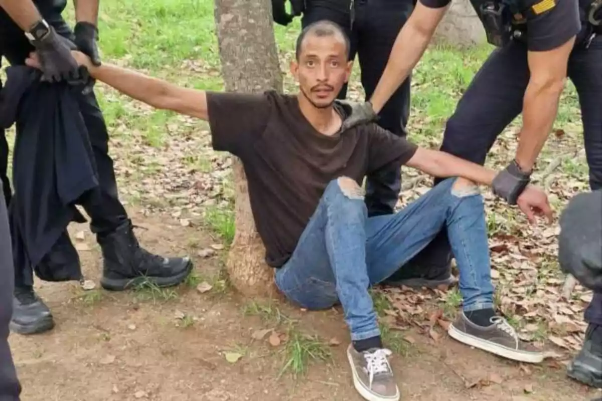 A man in a black T-shirt and ripped jeans sits on the ground leaning against a tree while being held by several people in dark uniforms.