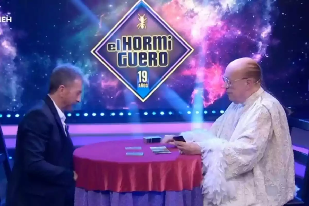 Pablo Motos i Rappel estan asseguts en una taula rodona coberta amb un mantell vermell en un plató de televisió amb un fons acolorit que mostra el logotip de "El Hormiguero 19 anys".