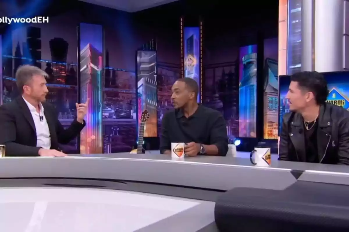 Pablo Motos, Anthony Mackie i Danny Ramírez asseguts a 'El Hormiguero', conversant al voltant d'una taula amb tasses de cafè, amb un fons de llums i edificis il·luminats.