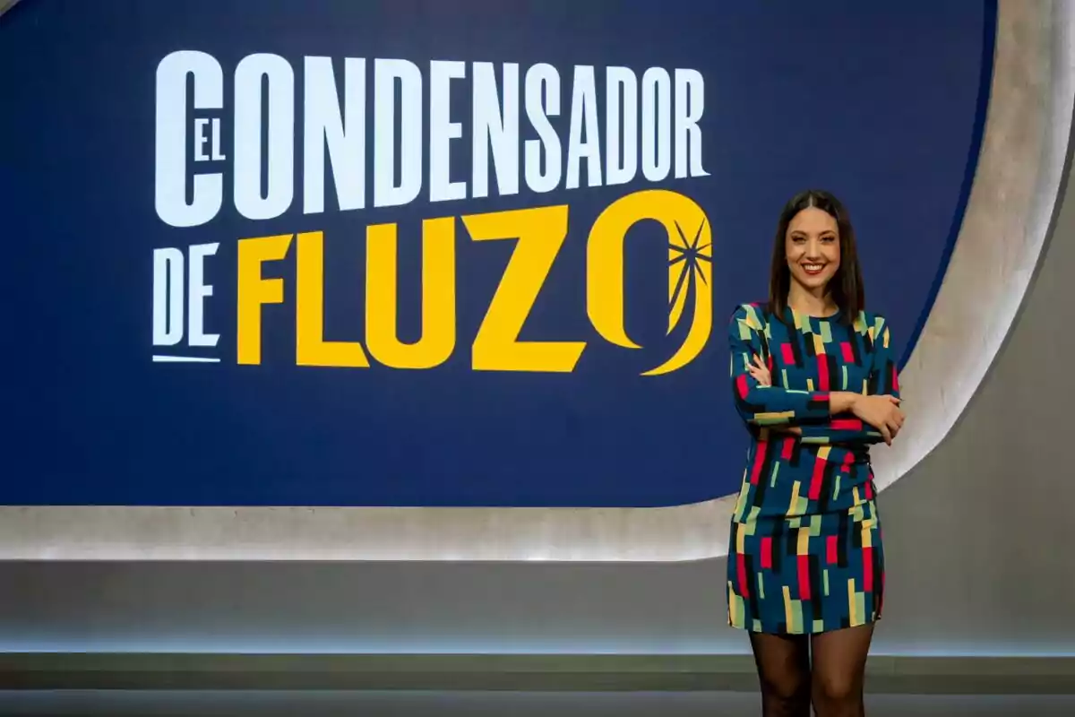 Maya Pixelskaya somrient amb un vestit colorit està dret davant d'un cartell que diu "El Condensador de Fluzo".