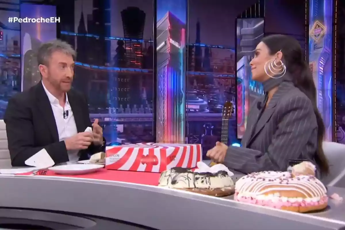 Pablo Motos i Cristina Pedroche conversen a 'El Hormiguero' amb un fons colorit i diversos pastissos sobre la taula.