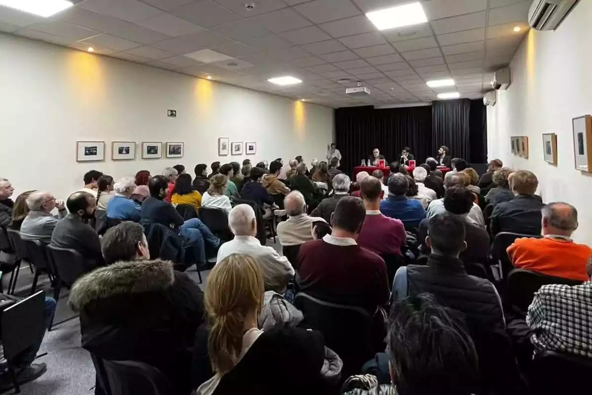 Desenes de persones, assegudes i d'esquena, segueixen un acte presentat per tres persones que presideixen la sala al fons de la imatge