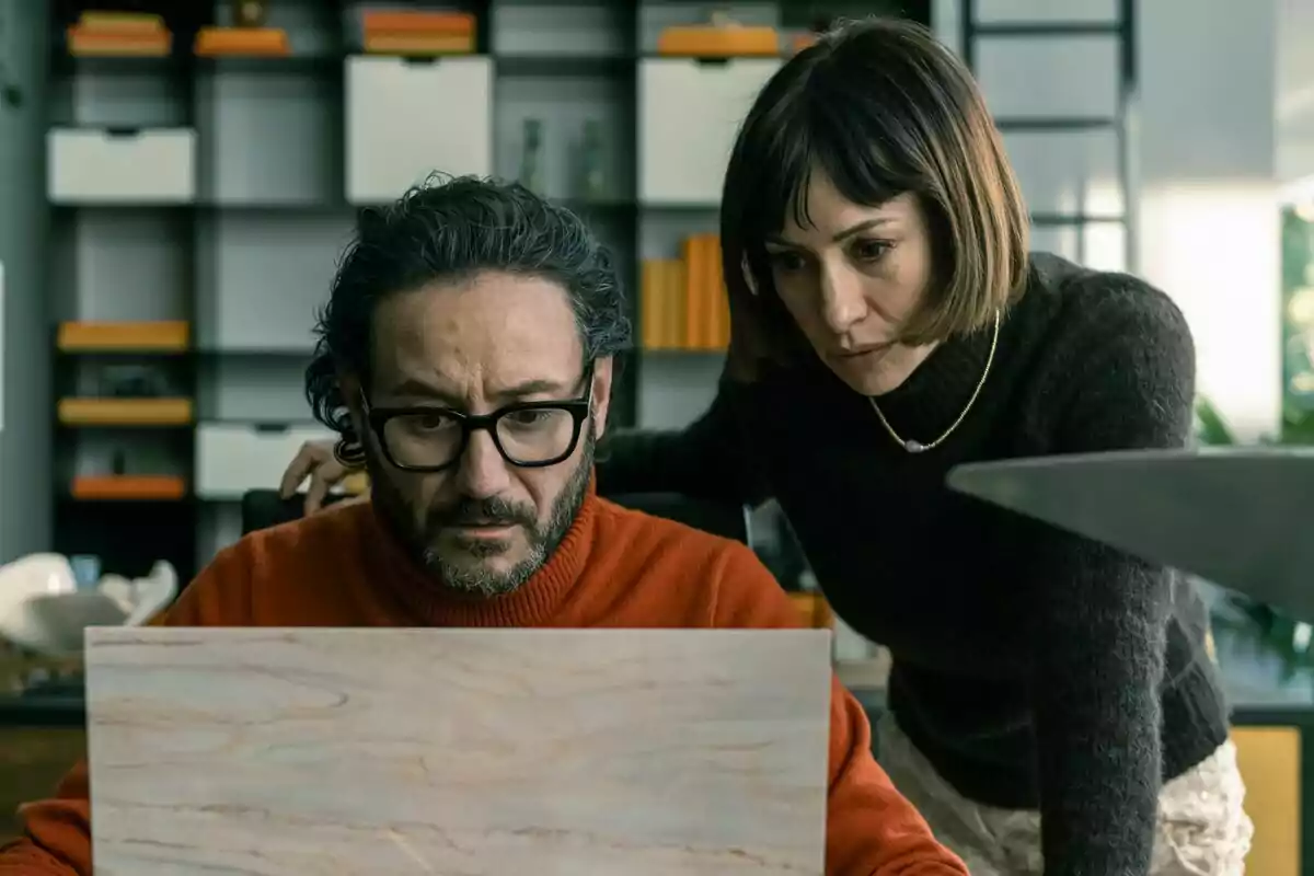 Carlos Santos amb ulleres i jersei taronja mira preocupat la pantalla d’un portàtil mentre una dona de cabell curt i jersei fosc l’observa atentament des del seu costat en una oficina moderna a El Refugi Atòmic. Carlos Santos amb ulleres i jersei taronja mira preocupat la pantalla d’un portàtil mentre una dona de cabell curt i jersei fosc l’observa atentament des del seu costat en una oficina moderna a El Refugi Atòmic.