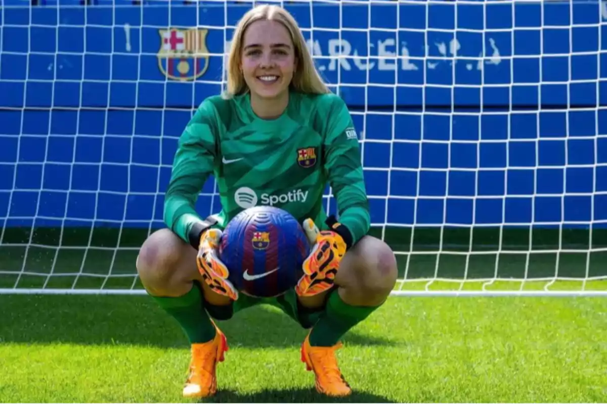 Portera del FC Barcelona femení somriu davant de la porteria mentre sosté una pilota amb totes dues mans i porta uniforme verd amb guants i botes taronges. Portera del FC Barcelona femení somriu davant de la porteria mentre sosté una pilota amb totes dues mans i porta uniforme verd amb guants i botes taronges.