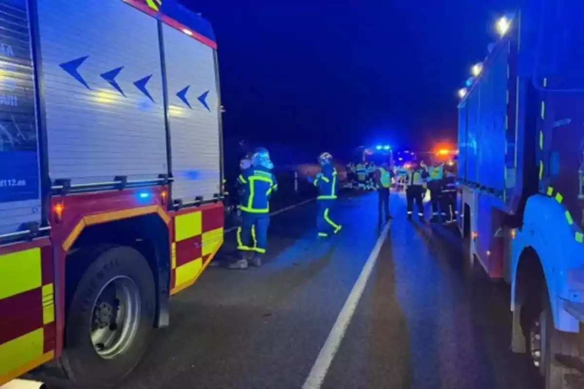 Camions de bombers i personal d'emergència treballant a una carretera a la nit amb llums blaves i grogues il·luminant l'escena.