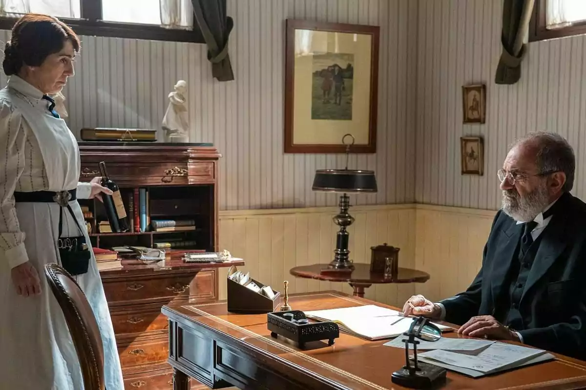 Arantxa Aranguren amb uniforme de servei sosté una ampolla de vi mentre parla amb Joaquín Climent més gran assegut en un escriptori en una oficina decorada amb mobles de fusta i quadres a les parets a La Promesa. Arantxa Aranguren amb uniforme de servei sosté una ampolla de vi mentre parla amb Joaquín Climent més gran assegut en un escriptori en una oficina decorada amb mobles de fusta i quadres a les parets a La Promesa.