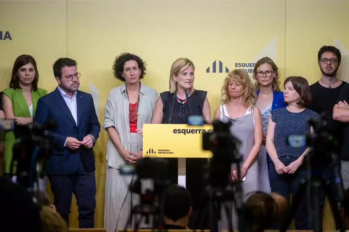 Un grup de persones dretes davant d'un podi groc amb el logotip d'Esquerra Republicana, mentre diverses càmeres els graven.