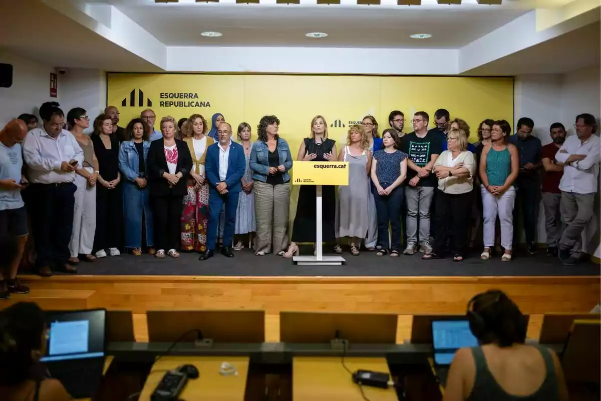 Un grup de persones dretes en un escenari amb un fons groc i un podi al centre.