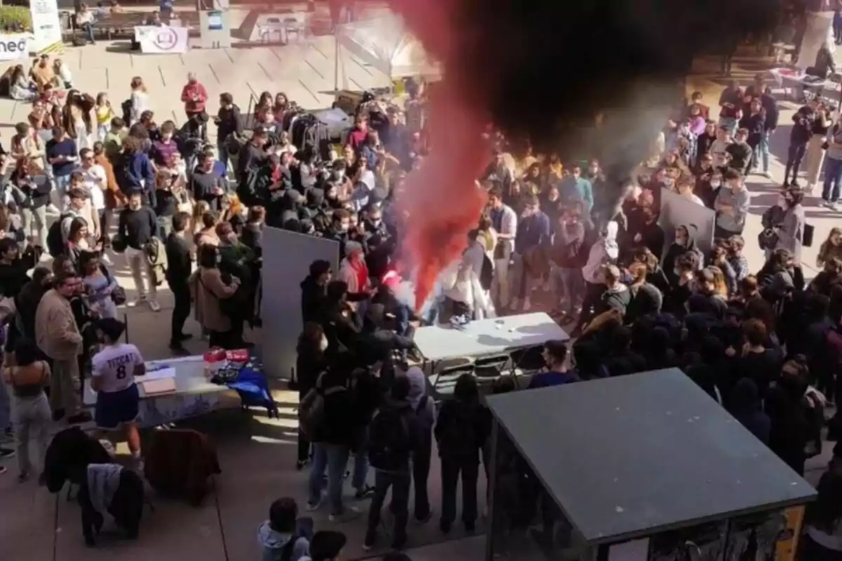 Un grup nombrós de persones es reuneix al voltant d’una taula mentre una columna de fum vermell i negre s’eleva al centre de l’esdeveniment a l’aire lliure.