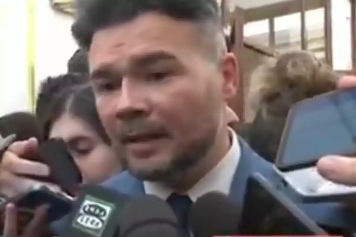 Home amb barba i vestit blau parlant amb la premsa envoltat de micròfons i telèfons mòbils