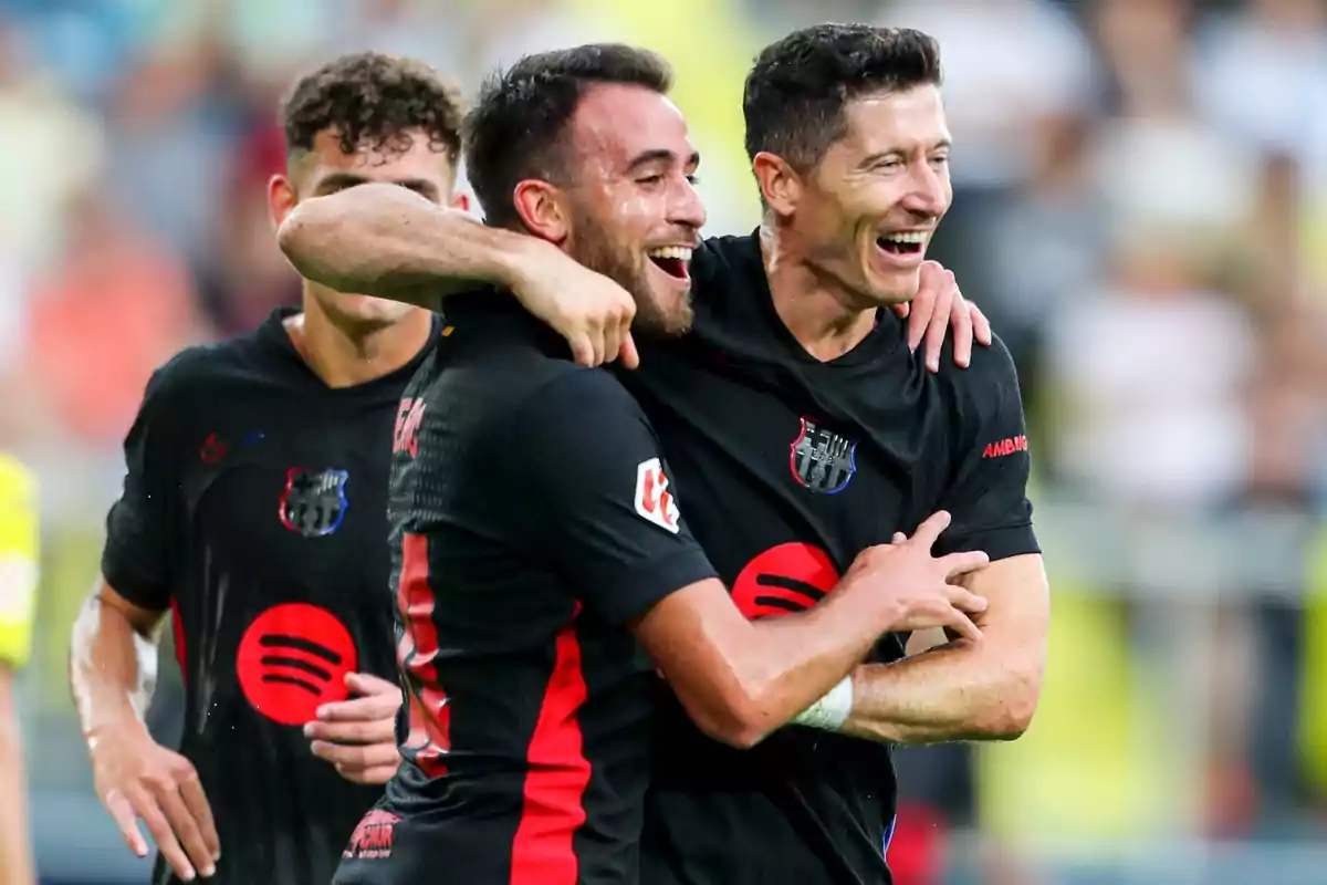 Jugadors de futbol celebrant un gol durant un partit, vestint uniformes negres amb detalls vermells i el logotip d'un patrocinador al pit.