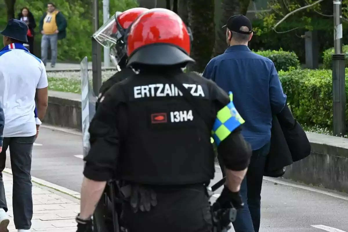 Agent de l’Ertzaintza amb casc vermell i uniforme negre caminant pel carrer al costat d’altres persones. Agent de l’Ertzaintza amb casc vermell i uniforme negre caminant pel carrer al costat d’altres persones.