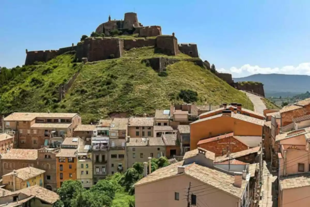 Una fortaleza histórica se alza sobre una colina verde con un pueblo de casas de techos de teja en primer plano bajo un cielo despejado.