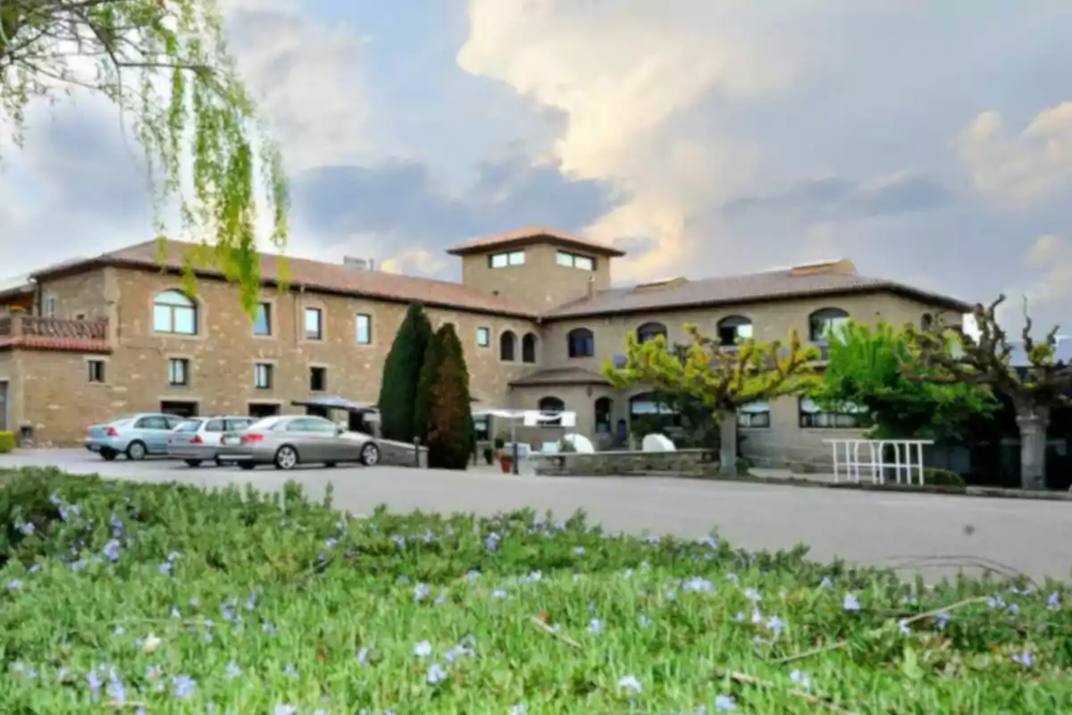 A stone building with a tiled roof surrounded by trees and shrubs, with several cars parked in front and a partially cloudy sky in the background.