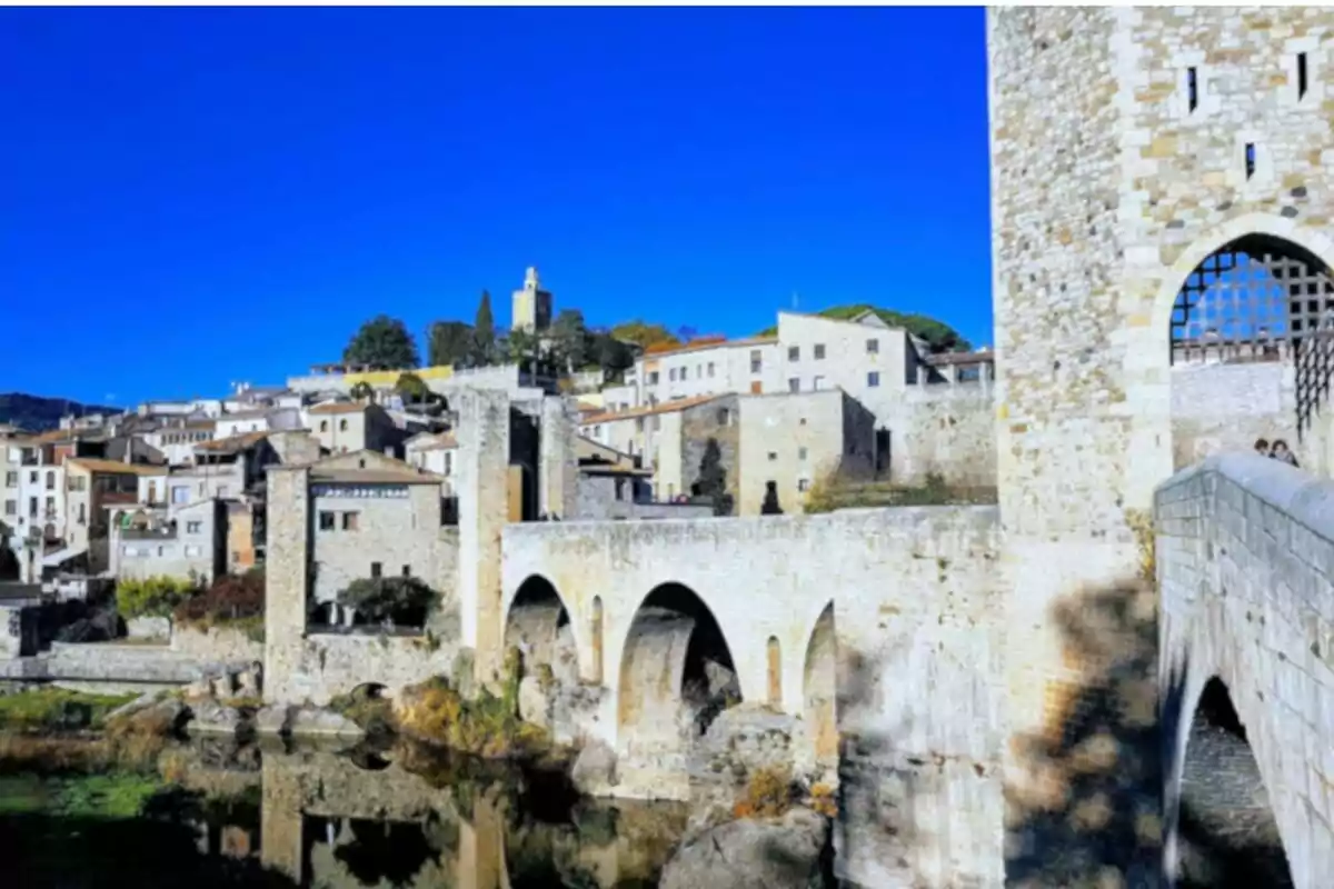 Vista de un puente medieval de piedra con arcos sobre un río y un pueblo histórico al fondo bajo un cielo azul brillante.