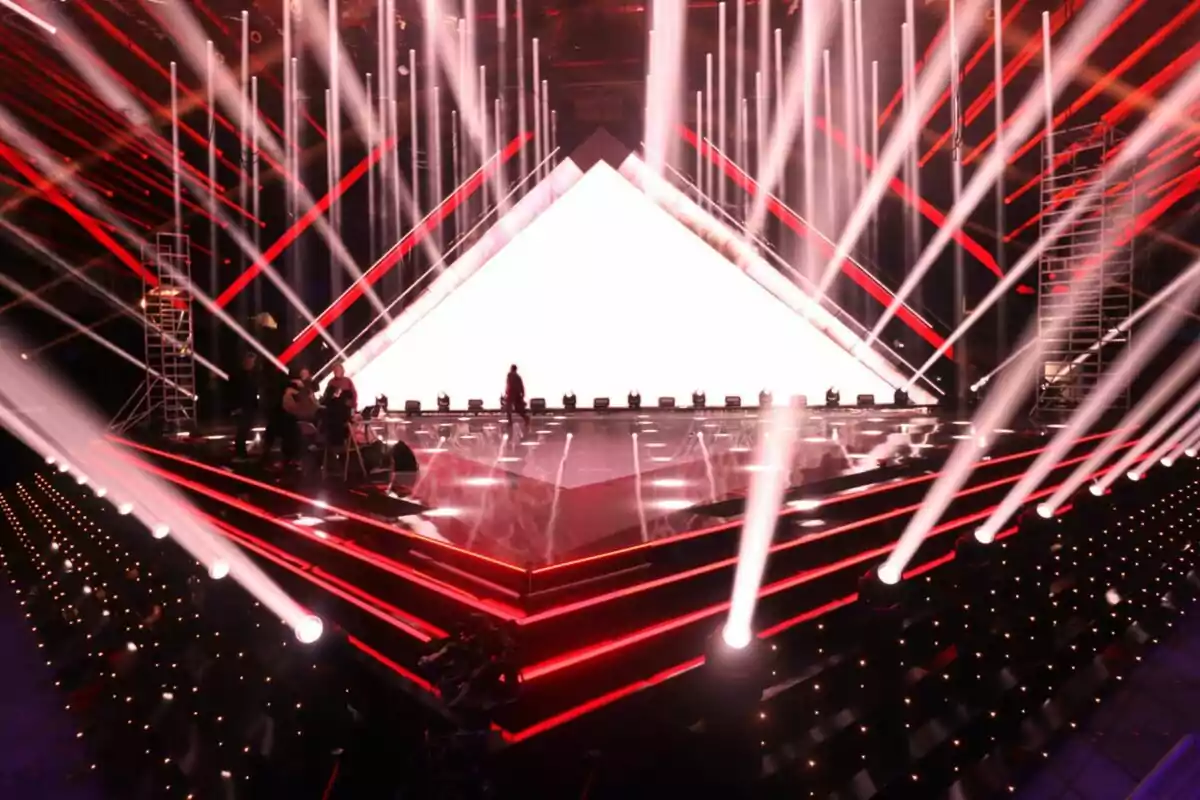 Un escenario iluminado del Benidorm Fest con luces rojas y blancas, con una estructura triangular brillante en el fondo y personas trabajando en el montaje.