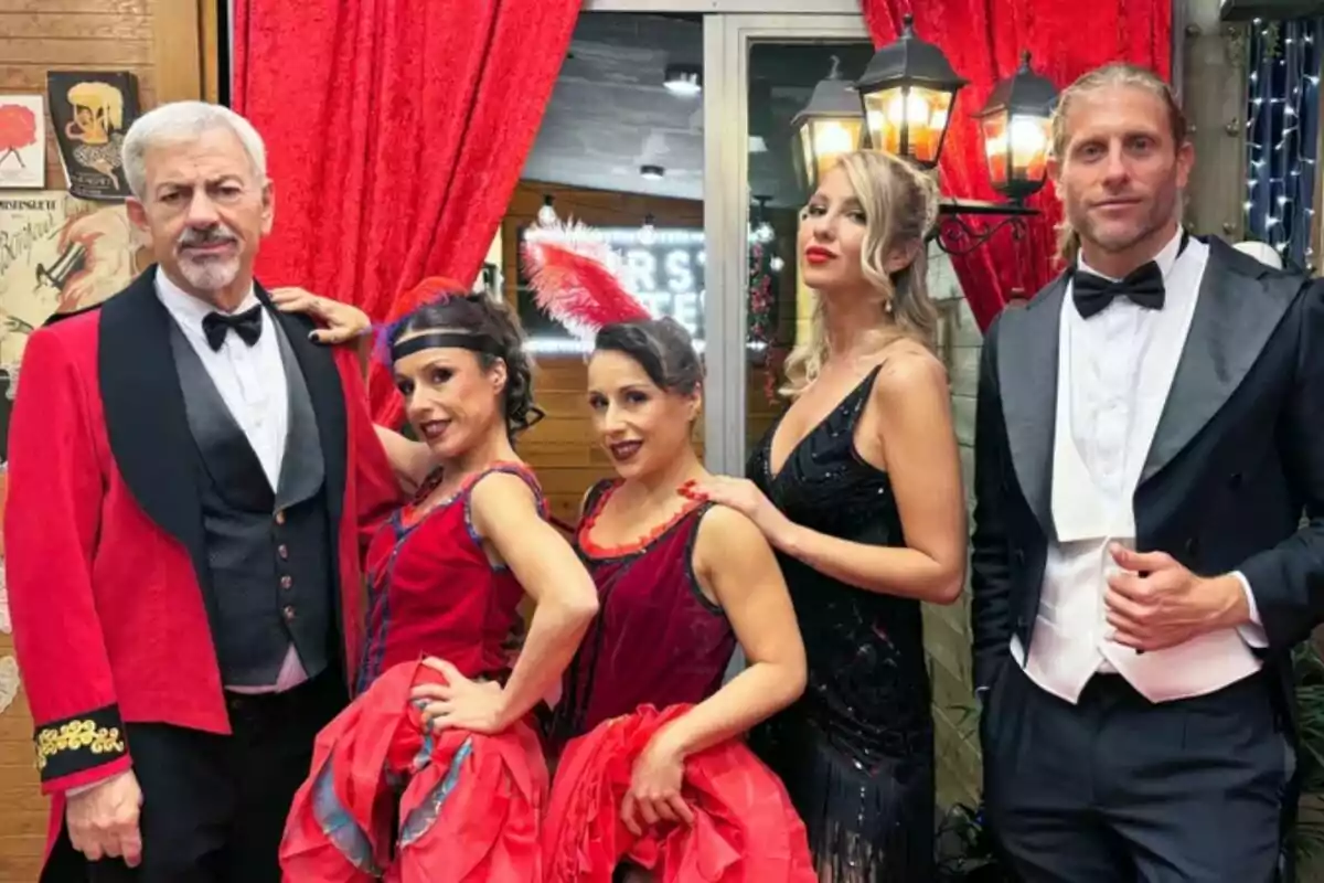 Un grupo de personas vestidas como Carlos Sobera con trajes elegantes y de época posan frente a un fondo decorado con cortinas rojas y luces en el especial de Nochevieja de First Dates.