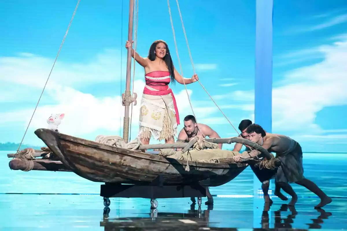 Esperansa Grasia disfressada de Moana està dreta en una balsa mentre tres homes l’empenyen en un escenari amb fons de mar i cel blau a Tu Cara Me Suena. Esperansa Grasia disfressada de Moana està dreta en una balsa mentre tres homes l’empenyen en un escenari amb fons de mar i cel blau a Tu Cara Me Suena.