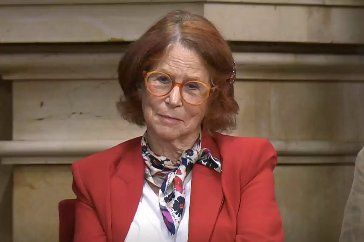 An older woman with orange glasses and brown hair, dressed in a red jacket and a patterned scarf, sits in front of a stone background.