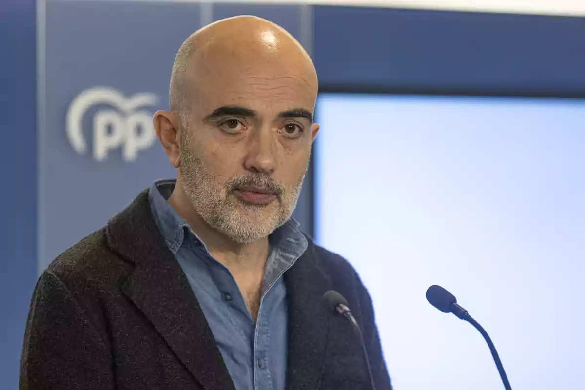 Home calb amb barba parlant a una conferència de premsa amb un fons blau i el logotip del PP.