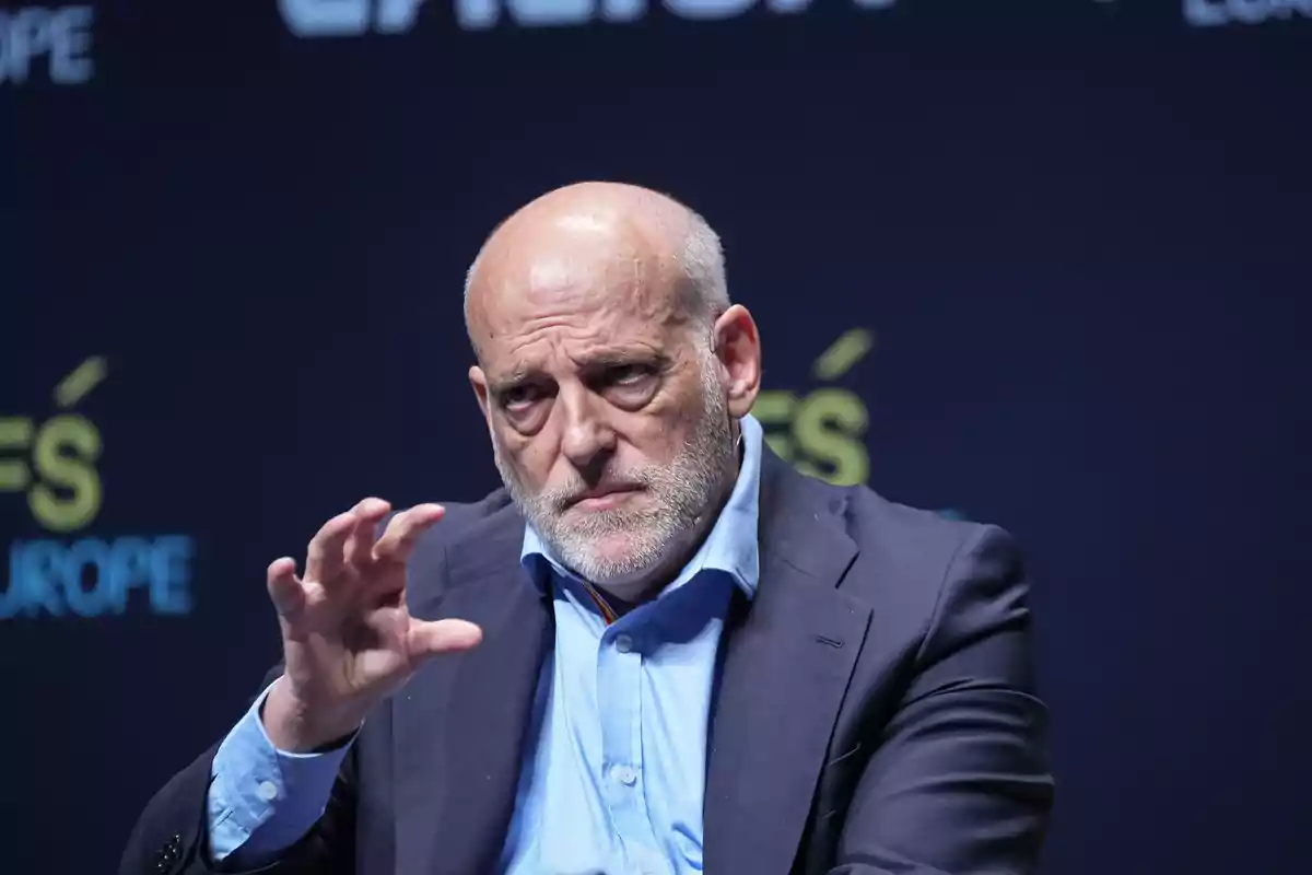 Bald man with beard and dark suit gesturing with hand in front of dark background with blurred text. Bald man with beard and dark suit gesturing with hand in front of dark background with blurred text.