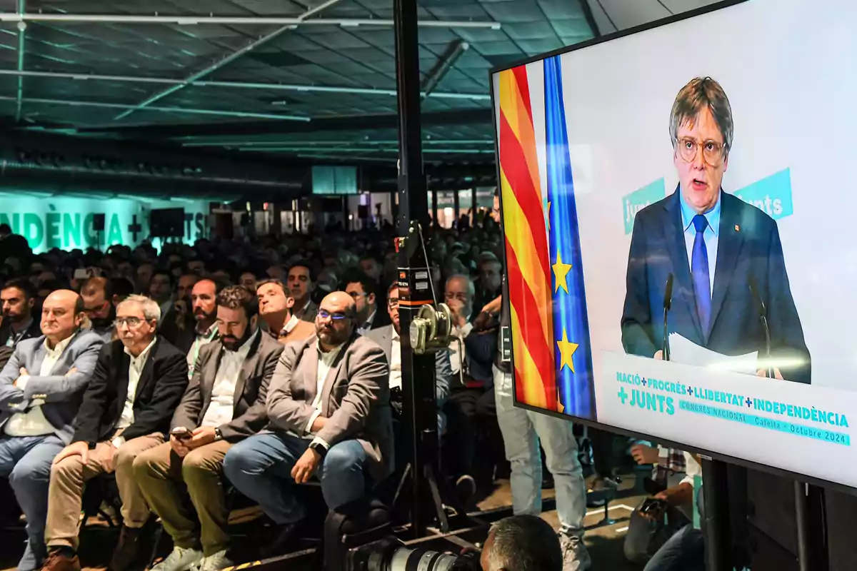 Una multitud de persones assegudes en un esdeveniment mentre observen una pantalla gran que mostra un orador amb banderes darrere seu.