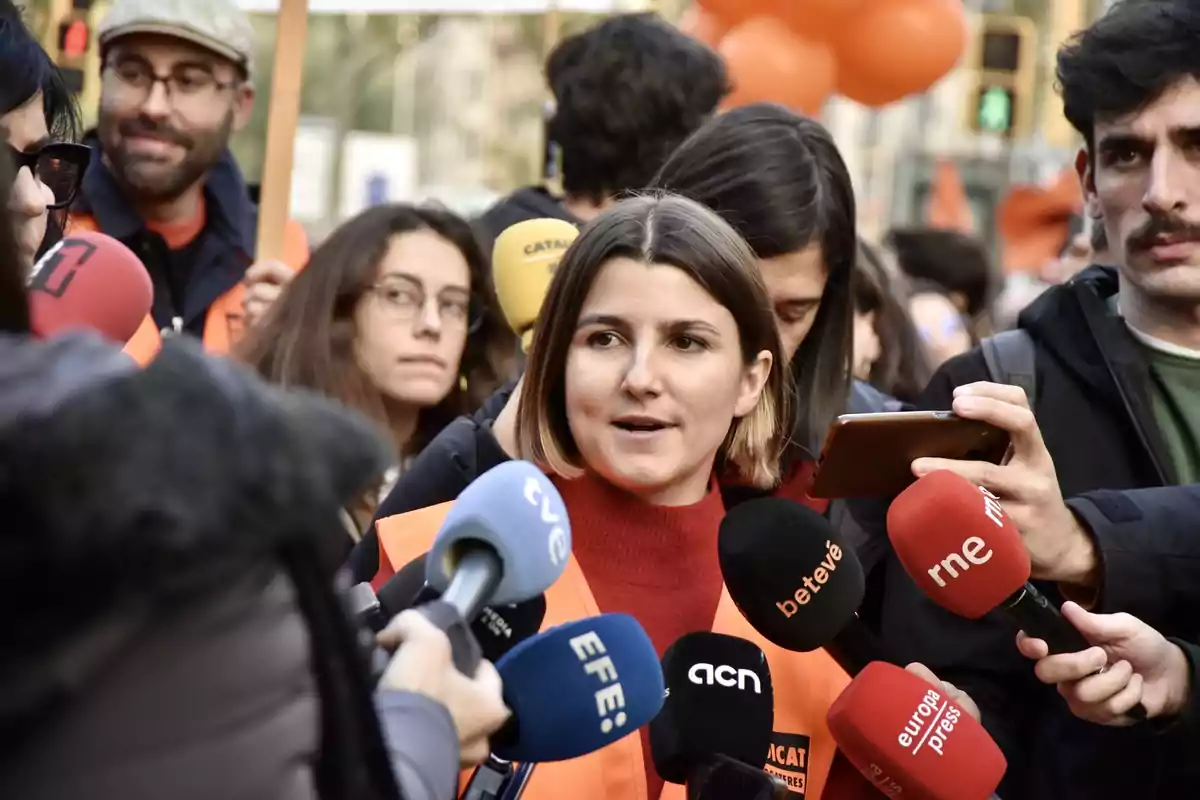 Una dona amb armilla taronja parla davant de diversos micròfons de diferents mitjans de comunicació envoltada de persones en un esdeveniment a l'aire lliure.