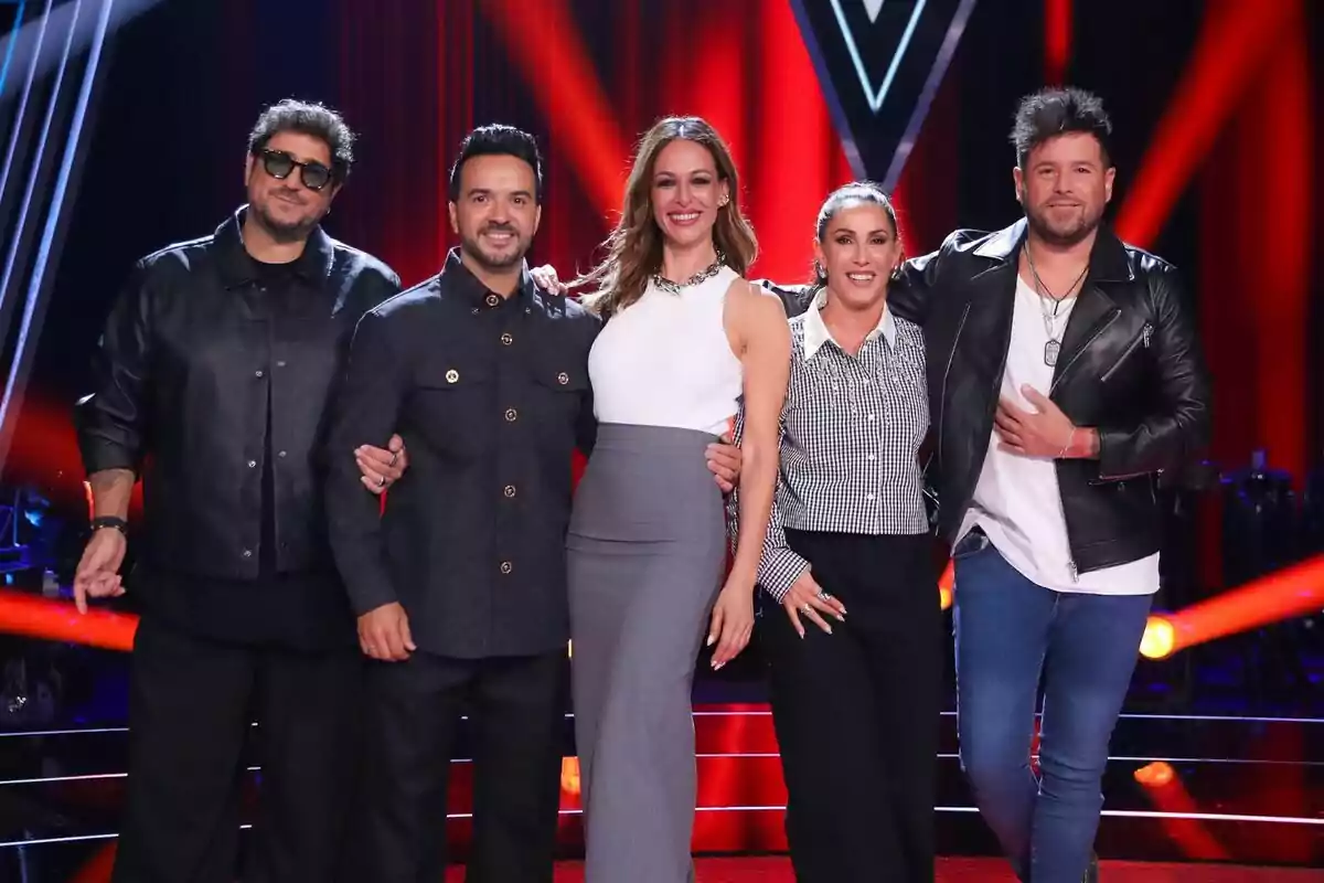 Eva González amb Pablo López, Antonio Orozco, Luis Fonsi i Malú posen juntes al plató de La Voz il·luminat amb llums vermelles i negres, amb un fons que inclou un logotip en forma de "V".