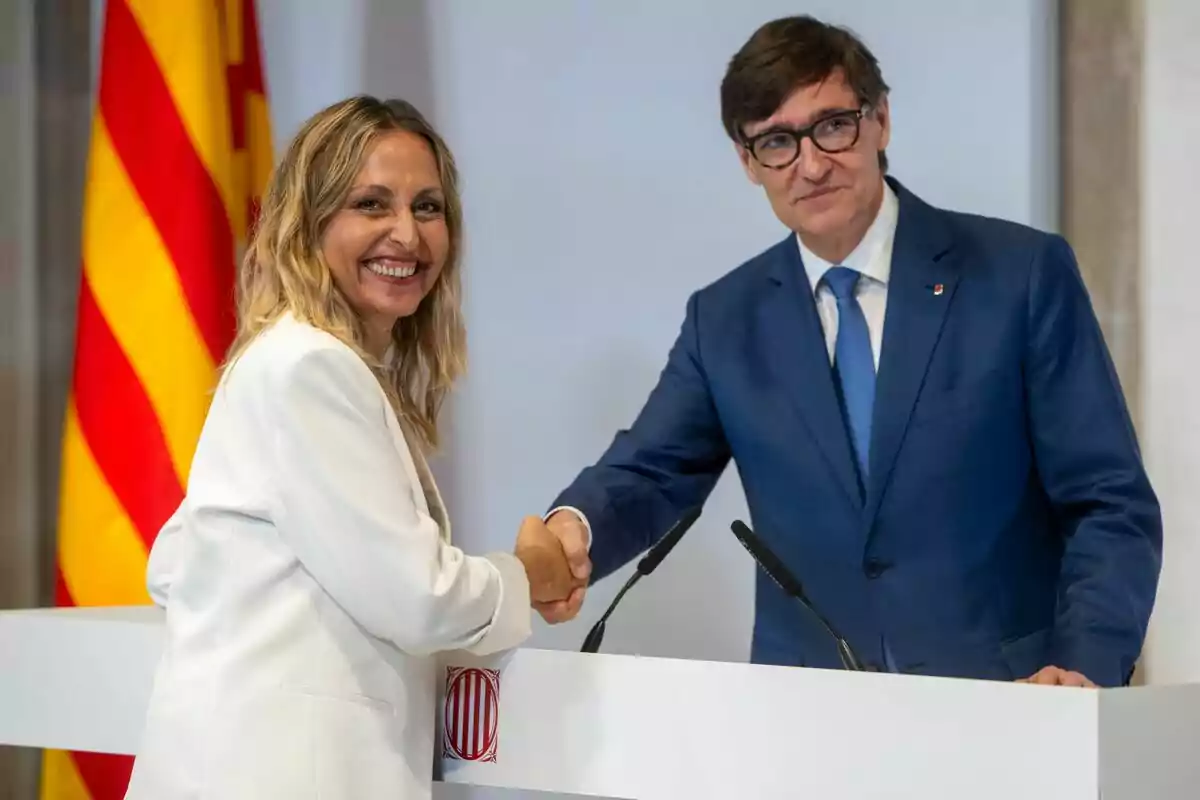 Eva Menor i Salvador Illa estrenyent-se la mà davant d'un podi amb una bandera catalana de fons.