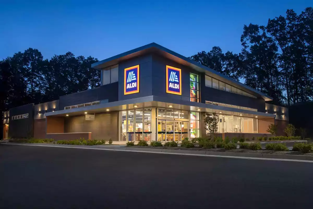 Fachada de una tienda ALDI iluminada al anochecer con árboles al fondo y un estacionamiento vacío al frente Fachada de una tienda ALDI iluminada al anochecer con árboles al fondo y un estacionamiento vacío al frente