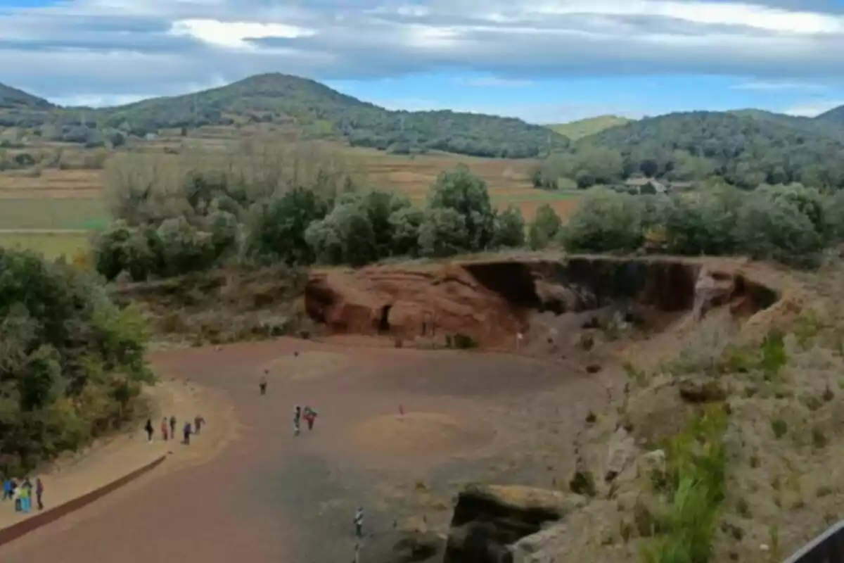 Un paisaje natural con un cráter rodeado de vegetación y colinas en el fondo, con varias personas caminando en el área central.