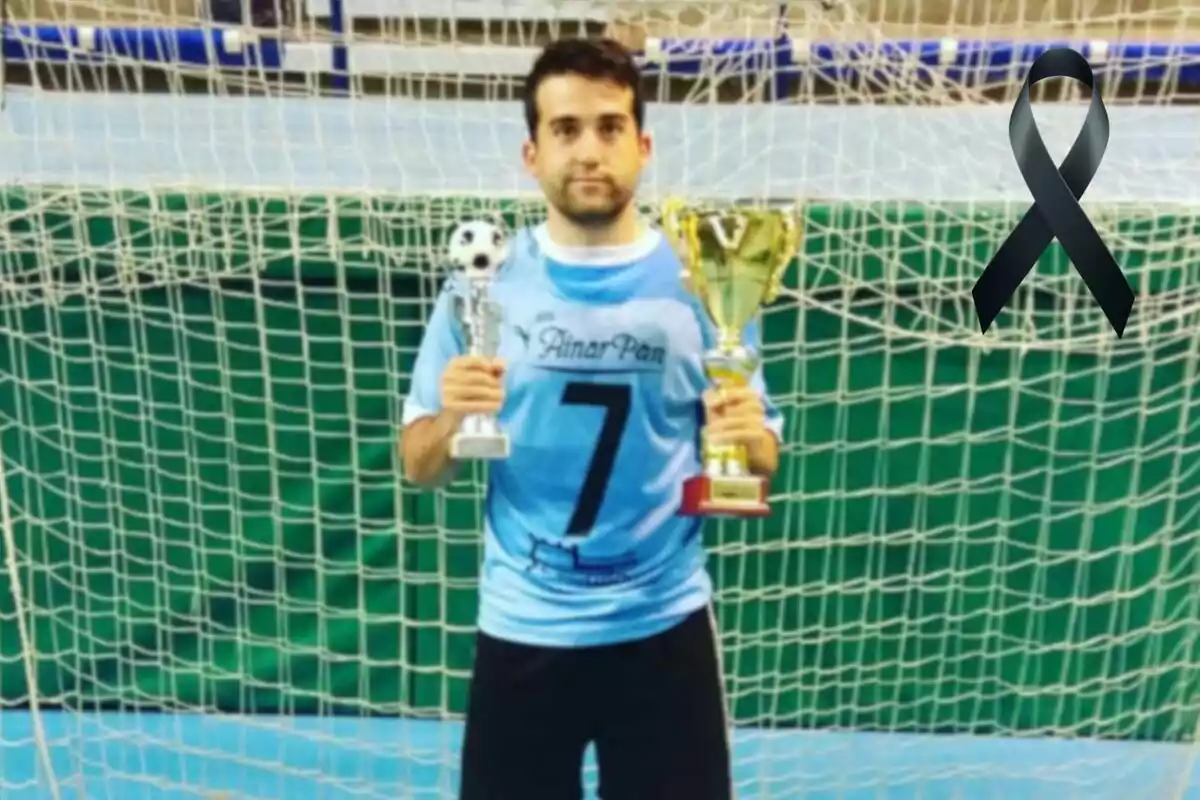 Un jugador de futbol sala amb samarreta blava i el número 7 sosté dos trofeus davant d'una porteria, amb un llaç negre a la cantonada superior dreta. Un jugador de futbol sala amb samarreta blava i el número 7 sosté dos trofeus davant d'una porteria, amb un llaç negre a la cantonada superior dreta.