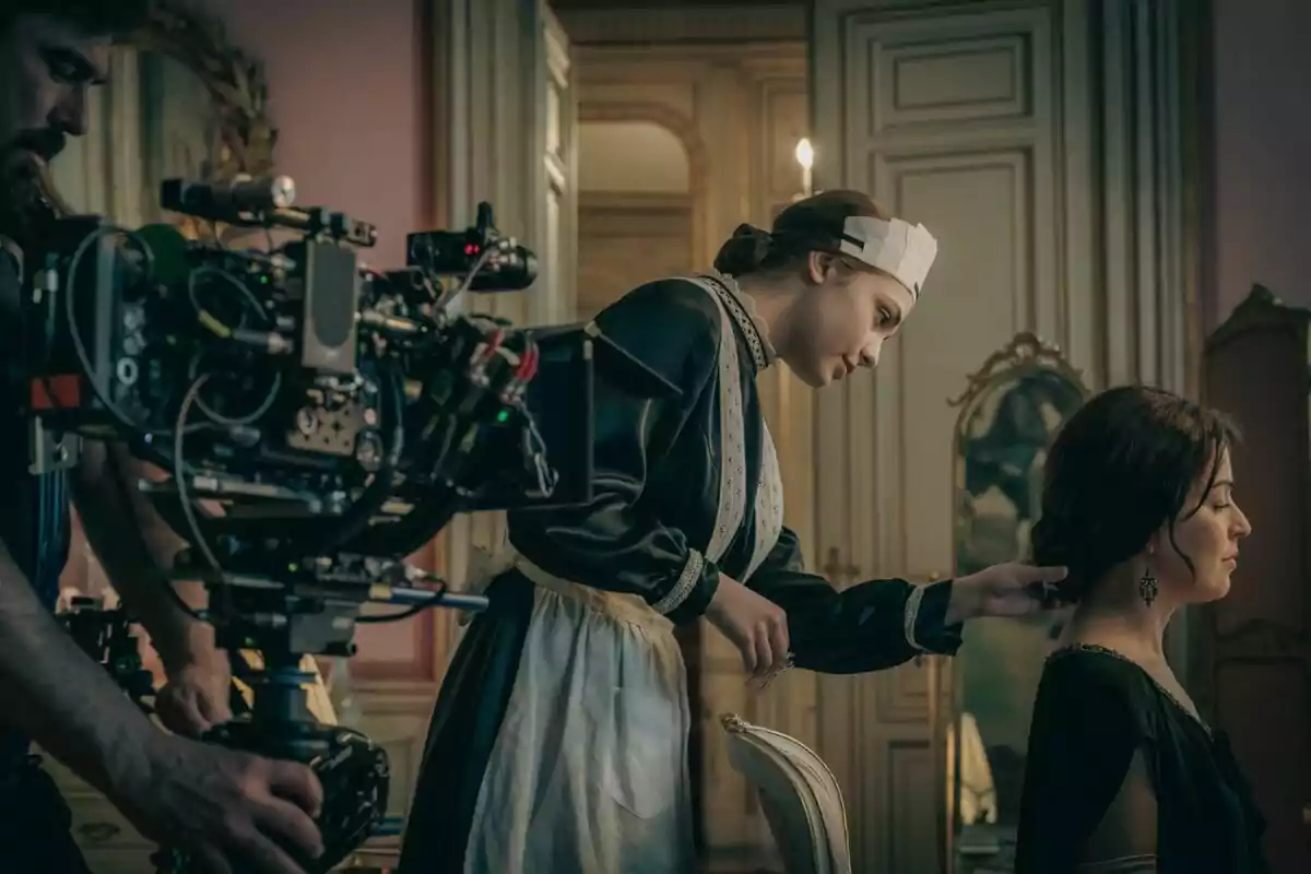 Una escena de rodatge mostra a Verónica Sánchez vestida d'època sent pentinada per una altra persona mentre un càmera grava l'acció en un elegant set. Una escena de rodatge mostra a Verónica Sánchez vestida d'època sent pentinada per una altra persona mentre un càmera grava l'acció en un elegant set.