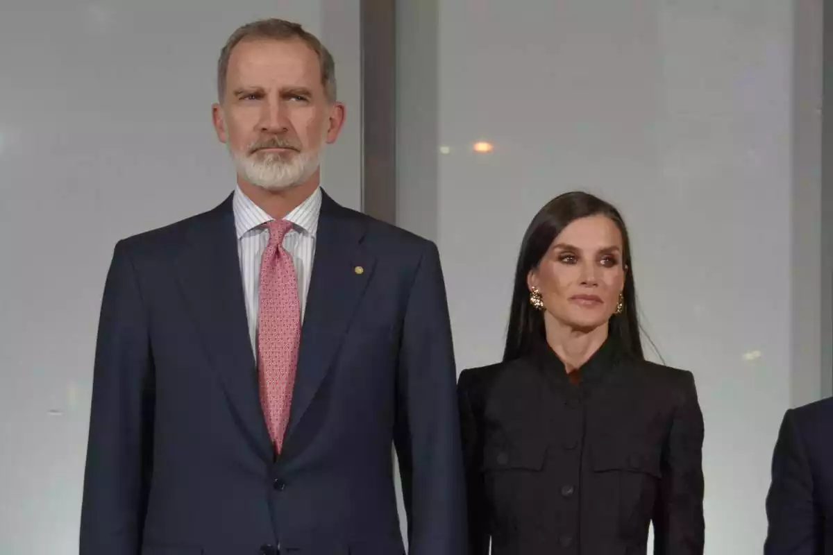 Felipe with a red tie and Letizia dressed in black serious looking to the side