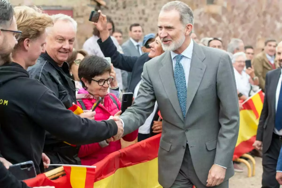 Felip VI amb vestit gris saluda persones darrere d’una tanca decorada amb banderes d’Espanya mentre diversos assistents fan fotos i somriuen. Felip VI amb vestit gris saluda persones darrere d’una tanca decorada amb banderes d’Espanya mentre diversos assistents fan fotos i somriuen.