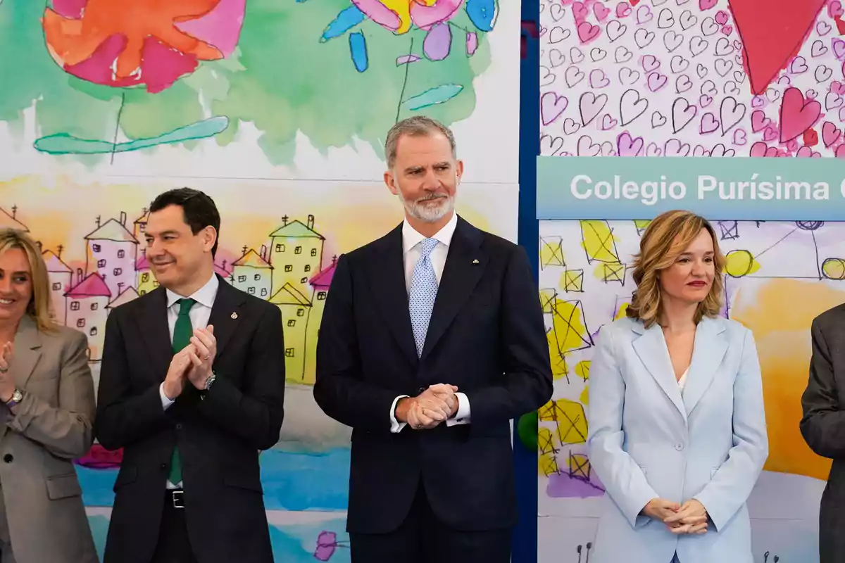 Several people dressed formally pose in front of a colorful background with children's drawings and a sign that says Colegio Purísima. Several people dressed formally pose in front of a colorful background with children's drawings and a sign that says Colegio Purísima.