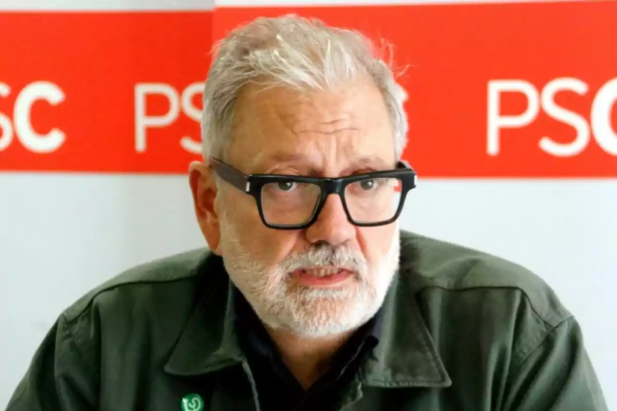 Un hombre con gafas y barba blanca está sentado frente a un fondo rojo con las letras "PSC".