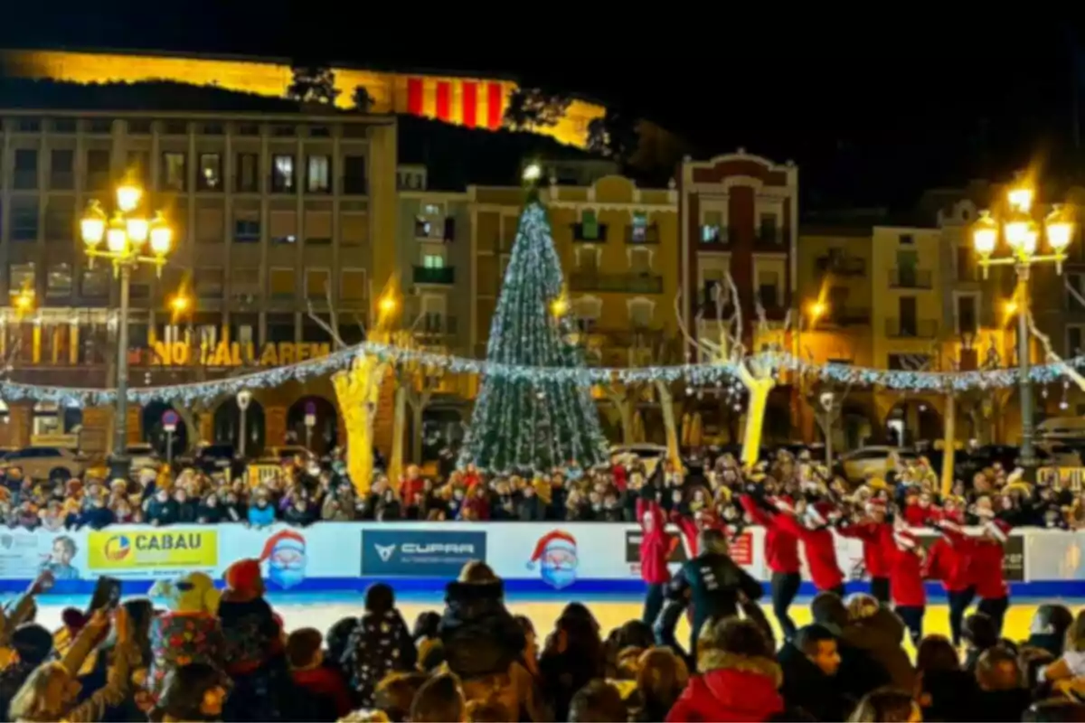 Una plaça il·luminada amb un arbre de Nadal i una pista de patinatge sobre gel envoltada de persones observant un espectacle.