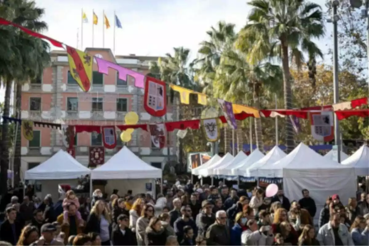 Una multitud gaudeix d'un esdeveniment exterior amb banderins colorits i botigues blanques, envoltats de palmeres i un edifici de fons.