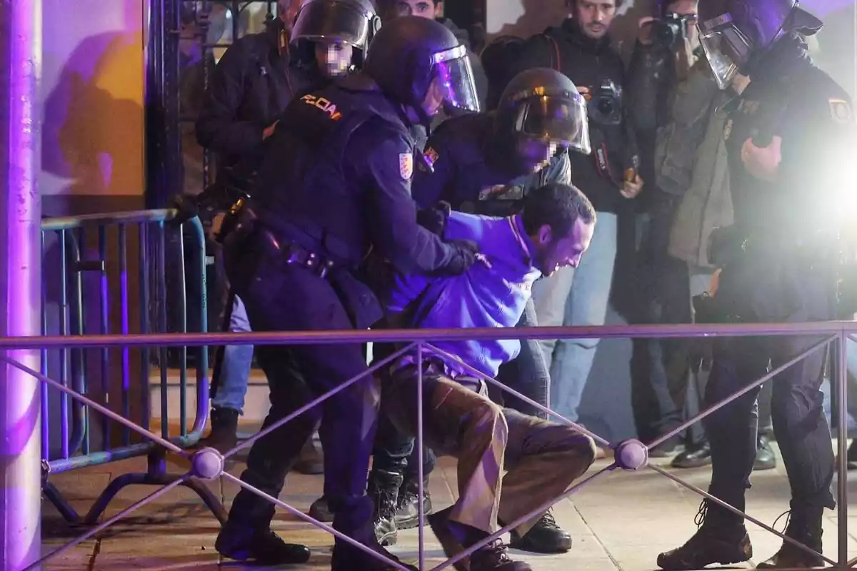 Dos agents de Policia antiavalots atura un manifestant al carrer Ferraz, davant de la seu del PSOE a Madrid, mentre dos agents més i diversos periodistes miren l'escena