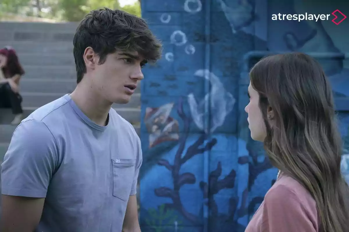 Dos personas conversan frente a un mural azul con motivos marinos mientras una tercera persona está sentada en las gradas al fondo en FoQ. La Nueva Generación.