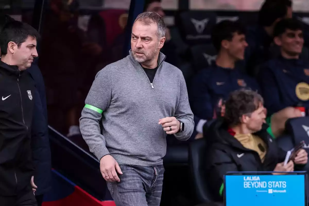 A man in a gray sweater and a green bracelet is standing next to a group of people sitting on a sports bench.