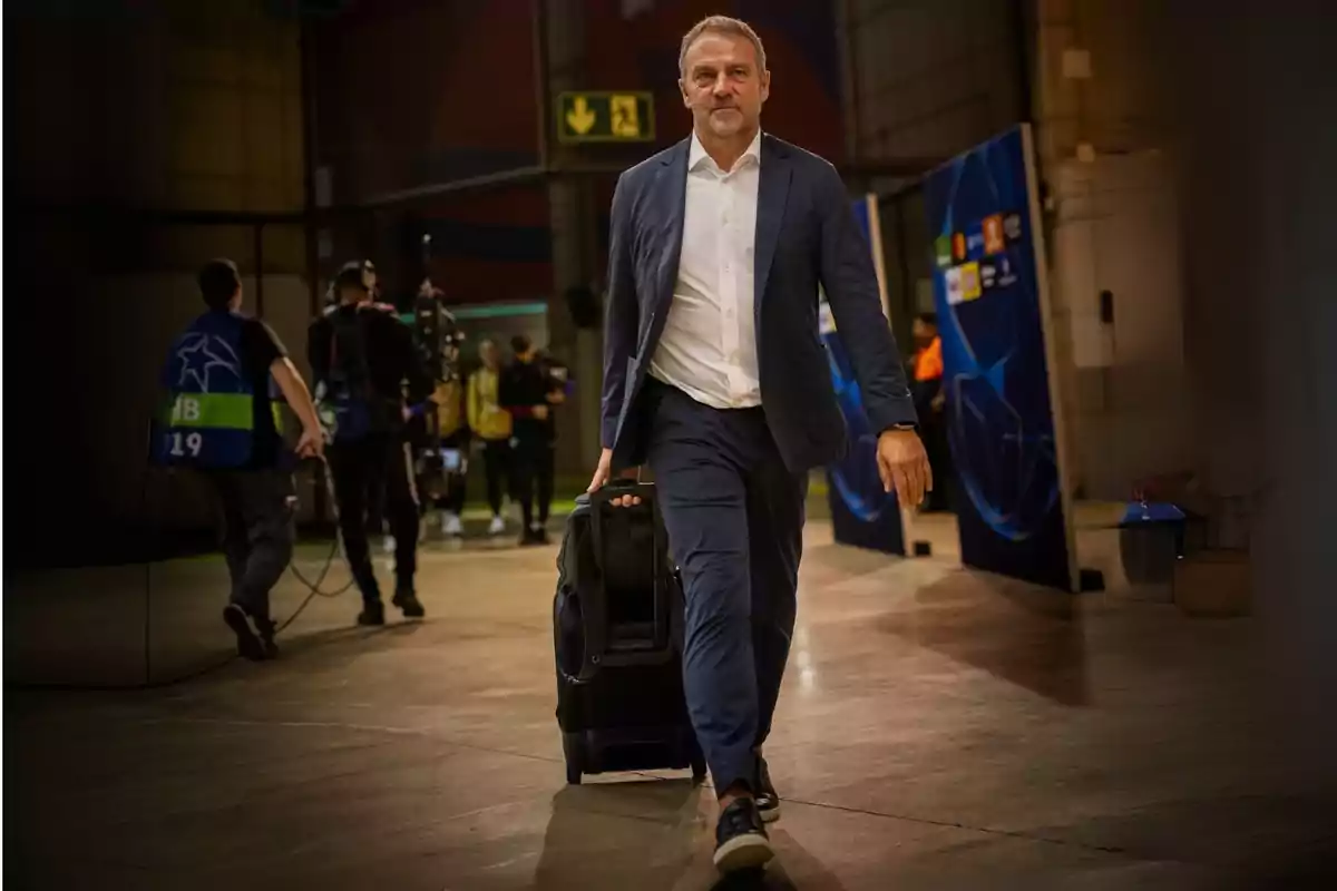 A man in a blue suit and white shirt walks down a hallway carrying a wheeled suitcase, while several people with filming equipment stand in the background.